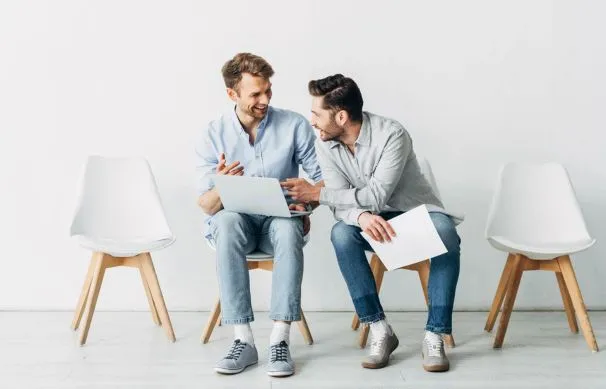 Two men sitting in chairs looking at a laptop discussing