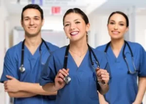 Three healthcare professionals in blue scrubs and stethoscopes smiling at the camera.